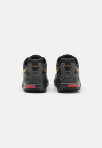 Sapatos atléticos pretos com um topo de rede texturizada, detalhes em fibra de carbono, um colar acolchoado e pormenores em vermelho no calcanhar e na sola.