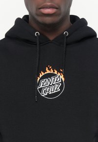 Zwarte hoodie met een rond geborduurd logo met "SANTA CRUZ" en vlammen in oranje. Inclusief koordjes en metalen oogjes.