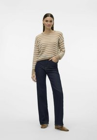 Pull en maille rayé beige et crème, associé à un jean large bleu foncé. Le pull présente une coupe ample et des poignets côtelés.