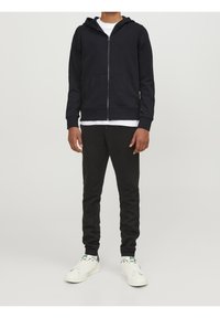 Marineblauwe zip-up hoodie met een voorzak, gecombineerd met zwarte sweatpants en witte sneakers met groene accenten. Eenvoudig textielontwerp, casual pasvorm.