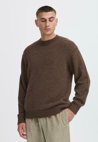 Brūns adīts džemperis ar klasisko kakla izgriezumu un ribotiem piedurkņu galiem. Mīksta tekstūra, brīvs piegriezums, saskaņots ar gaišas krāsas korduroja biksēm.