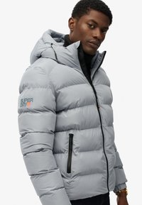 Giubbotto imbottito grigio chiaro con cappuccio, dotato di chiusura con zip, due tasche laterali con zip e logo "Superdry" sulla manica sinistra.
