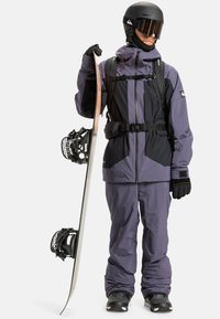 Fialovo-čierny snowboardový outfit s nepremokavým materiálom, zateplenou bundou, lyžiarskymi okuliarmi, rukavicami a snowboardom s viazaniami.