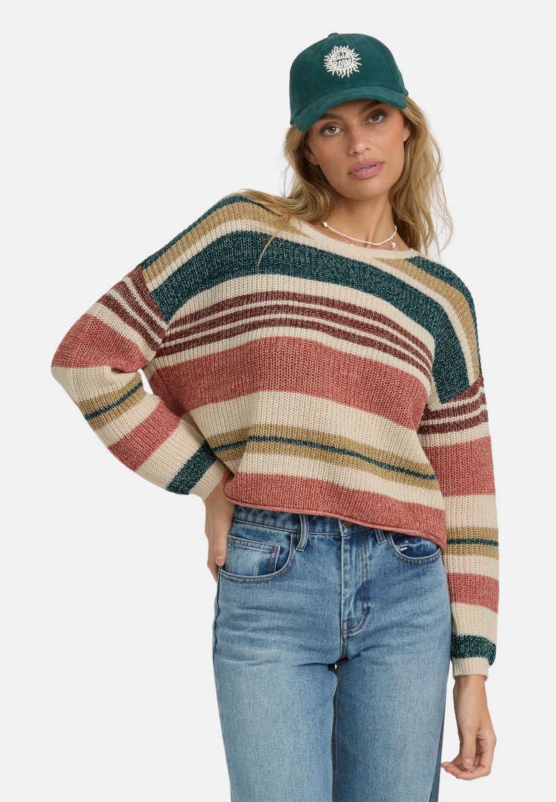 Maglione a righe in crema, ruggine e verde; vestibilità morbida con maniche larghe. Abbinato a jeans blu a vita alta e un cappellino verde.