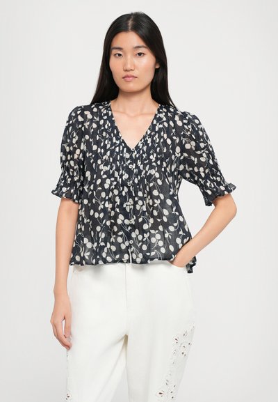 Bluză în albastru închis cu model floral alb, decolteu în V, detalii cu nasturi, mâneci scurte și puff, dintr-un material ușor și texturat. Pur tată cu pantaloni albi.