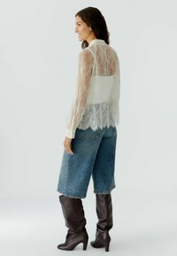 Doorzichtige kanten blouse met lange mouwen, crème kleur, gelaagd over een effen witte top; gecombineerd met hoge denim shorts met hoge taille en bruine kniehoge laarzen.