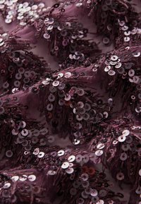 Tissu bordeaux recouvert de paillettes métalliques, agencées en grappes, créant une surface texturée et scintillante avec des franges délicates.
