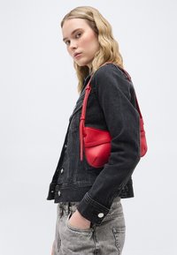 Νέα γυναίκα με ξανθά κυματιστά μαλλιά που φοράει μαύρο μπουφάν από τζιν και γκρι τζιν, κρατώντας μια μικρή κόκκινη δερμάτινη τσάντα στον ώμο.
