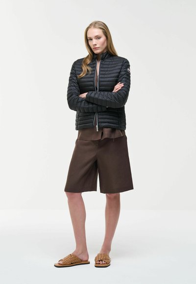 Femme debout, les bras croisés, portant une veste matelassée noire, un short marron arrivant aux genoux, et des sandales tissées de couleur beige sur un fond uni.