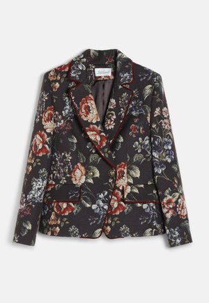 Donna che indossa un blazer floreale e scarpe coordinate, pantaloni larghi scuri, che tiene in mano una borsa nera con chiusura dorata, in piedi su uno sfondo semplice.