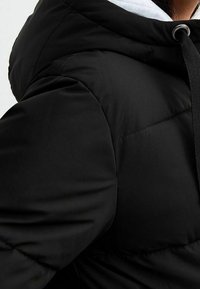 Musta toppatakki, jossa tikattu kuosi, pehmeä koostumus ja kiristysnauhalla varustettu huppu. Paljas vetoketju ja kontrastivärinen valkoinen vuori.