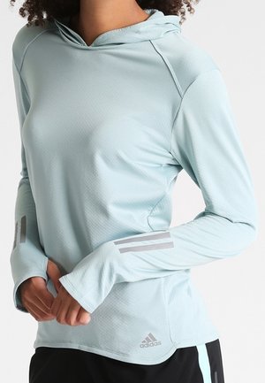 Kvinde iført lyseblå Adidas hoodie med lange ærmer, tommelfingerhuller og reflekterende striber på ærmerne, kombineret med sorte og blå bukser.