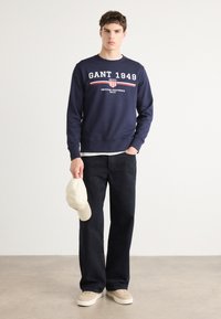 Marineblauwe sweatshirt met witte "GANT 1949" tekst en rode accenten, gecombineerd met donkere spijkerbroek, crèmekleurige pet en beige sneakers.