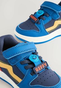 Niebieskie trampki z rzepami mają na sobie wyraźnie widoczne uśmiechnięte buźki oraz słowo "SZCZĘŚLIWY" na każdej z butów. Kolorowe elementy wzornicze zwiększają atrakcyjność.