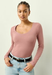 Langärmliges Oberteil mit U-Ausschnitt aus weichem pinken Stoff, kombiniert mit hellblauen High-Waist-Jeans und einem schwarzen Gürtel. Glatte Textur und figurbetontes Design.