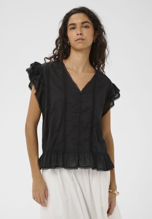 Mujer con blusa negra abotonada con mangas y dobladillo con volantes, combinada con falda blanca, de pie frente a un fondo liso.