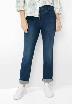 Kvinde iført mørkeblå cuffede jeans, hvid blomstret skjorte og hvide sneakers, stående mod en ensfarvet hvid baggrund.