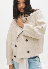 Giacca doppiopetto in misto lana beige con ampi rever, bottoni neri e una vestibilità rilassata. Presenta dettagli cuciti e tessuto testurizzato.