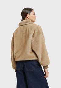 Giacca in pelliccia sintetica beige con ampio colletto, vestibilità comoda e polsini a costine. Indossata con pantaloni in denim blu scuro.