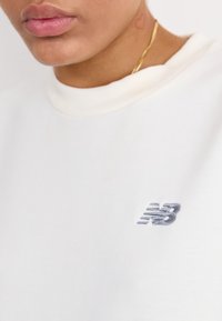 Sudadera blanca hecha de tela suave, con un pequeño logo gris bordado en el pecho y un cuello de canalé.