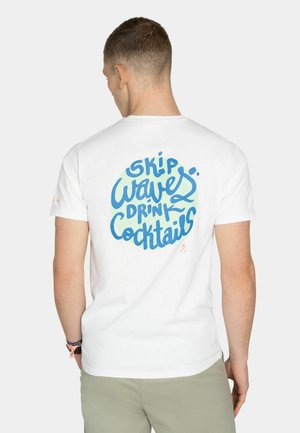 Hombre con camiseta blanca con texto azul "Skip Waves Drink Cocktails" en la parte trasera, pantalones gris claro y pulseras, de espaldas.