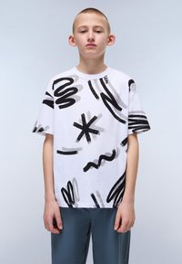 Weißes T-Shirt mit abstrakten schwarzen und grauen Pinselstrichmustern. Kurze Ärmel und runder Ausschnitt. Kombiniert mit grauen Hosen.