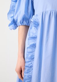Lyseblå kjole med voluminøse, flagrende ærmer og detaljer med flæser langs siden. Tekstiler med blød glans.