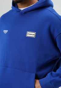 Blauwe katoenen hoodie met een voorzak, geribde manchetten en twee logo's: "STRONGER" en "chance" op de borst. Gladde textuur.
