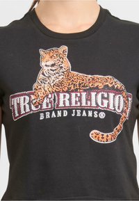 Czarna koszulka z wzorem lamparta ozdobionym cyrkoniami, obok której znajduje się nazwa marki "TRUE RELIGION" w pogrubionej czcionce.