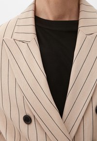 Blazer doppiopetto beige con righe verticali nere; presenta una texture liscia e due bottoni neri. Indossato su una camicia nera.