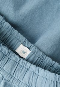 Lichtblauwe stof met een textuurafwerking, met een elastische tailleband en een wit label met een klein blauw logo. Close-up weergave.