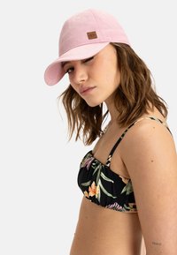 Gorra de algodón rosa claro con visera curva, que presenta un pequeño parche de logo marrón. El modelo lleva puesto un top de bikini negro con estampado floral.