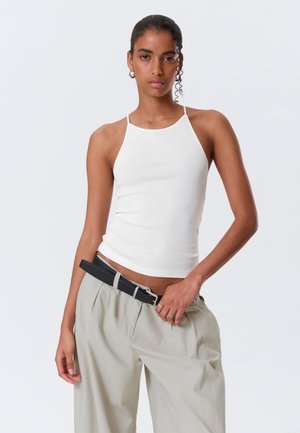 Jeune femme portant un haut blanc sans manches et un pantalon beige à jambes larges avec une ceinture noire, debout devant un fond clair uni.