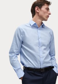 Helles blaues Langarmhemd mit Button-Down-Kragen und weißen Knöpfen, getragen mit navyblauen Hosen und einem schwarzen Gürtel. Glatte Textur, figurbetonter Schnitt.