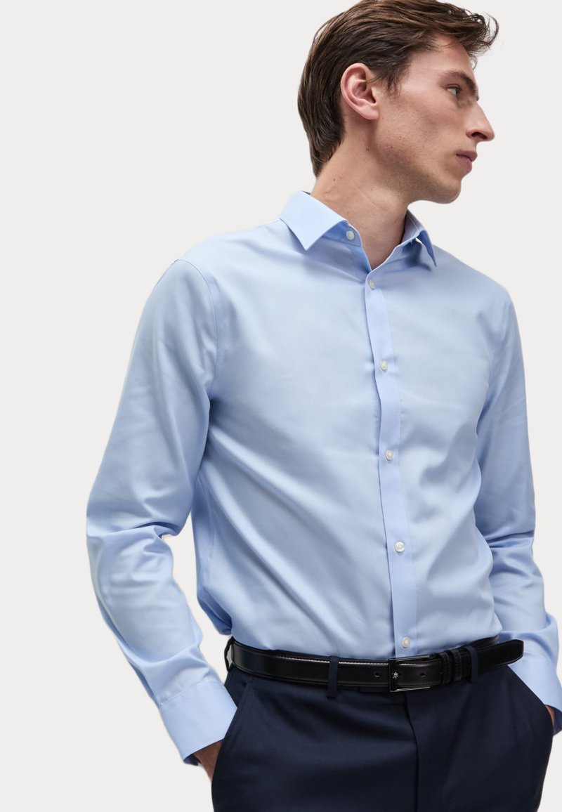 Helles blaues Langarmhemd mit Button-Down-Kragen und weißen Knöpfen, getragen mit navyblauen Hosen und einem schwarzen Gürtel. Glatte Textur, figurbetonter Schnitt.
