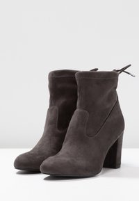 Botas de tobillo de ante gris con una textura suave, puntera redondeada y un tacón grueso. Presentan un diseño recortado sutil y un detalle de lazo en la parte trasera.