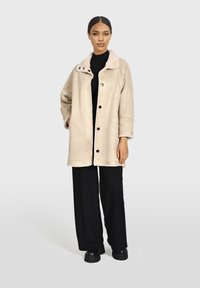 Abrigo de borrego beige con un suave cuello, botones frontales y bolsillos laterales. El modelo lleva pantalones negros y un suéter de cuello alto.