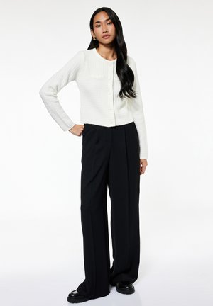 Vrouw staat met één hand op de heup, draagt een wit gebreid vest met knopen, zwarte wijde broek en zwarte schoenen tegen een witte achtergrond.