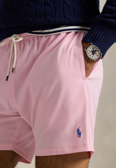 Oseba, ki nosi roza kratke hlače z belim vrvico in modrim vezenim logotipom, temno moder pulover in zapestno uro z motivom lika na številčnici.