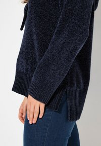Maglione lavorato a maglia blu navy con una finitura testurizzata, polsini a coste e fessure laterali. Indossato su jeans scuri, con cappuccio regolabile.