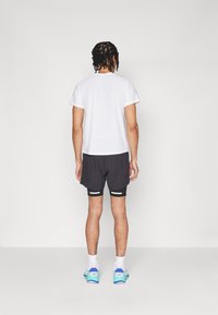 Weißes T-Shirt mit kurzen Ärmeln in Kombination mit schwarzen Sportshorts, die ein geschichtetes Design und Seitentaschen aufweisen. Blaue und grüne Laufschuhe sind sichtbar.