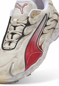 Beige sneakers med et tekstureret stofoverdel, røde og sorte accenter samt et karakteristisk bølget design. Har hvide snørebånd og en gummisål.