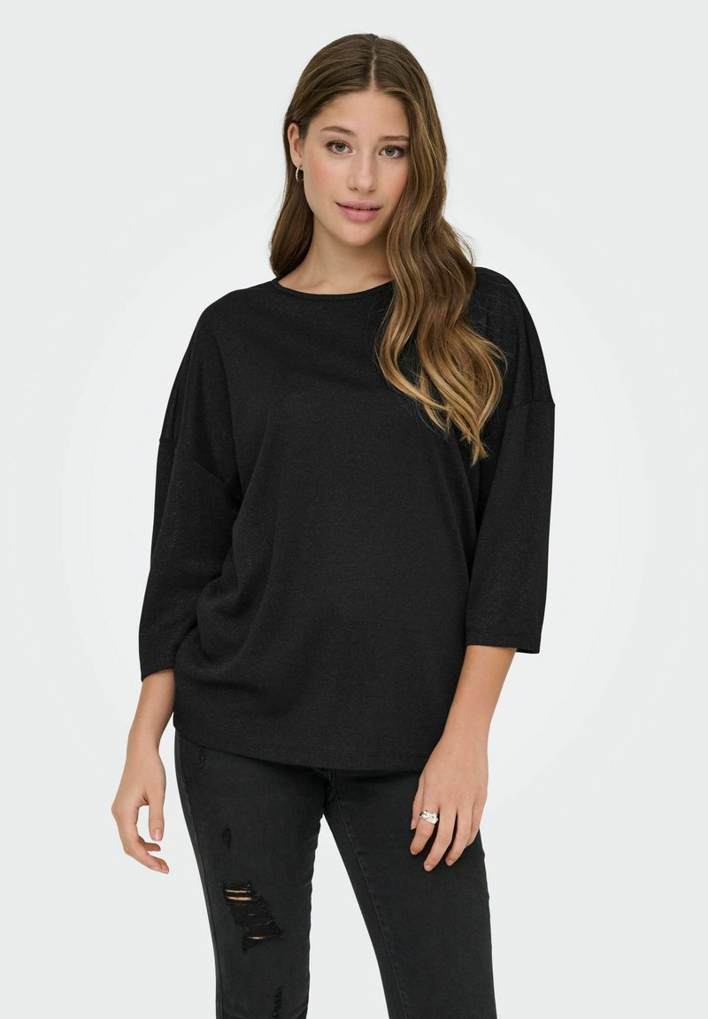 Joven mujer con cabello largo y ondulado, vestida con una blusa negra holgada de mangas tres cuartos y jeans negros rotos, de pie contra un fondo claro.