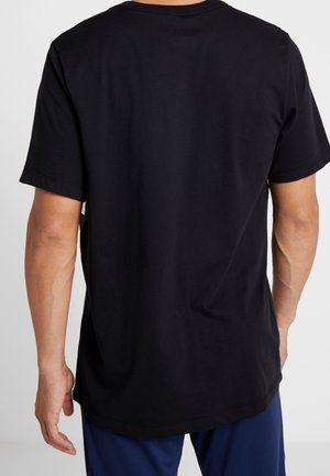 Mand iført en ensfarvet sort T-shirt med korte ærmer vist bagfra, kombineret med mørkeblå bukser mod en neutral baggrund.