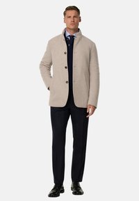 Gai bēša vilnas maisījuma mētelis ar augstu apkakli, priekšējo pogu un sānu kabatām, uzvilkts virs tumša džempera un līniju biksēm.