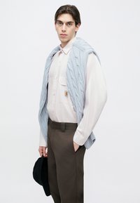 Ανοιχτό γαλάζιο πουλόβερ με πλέξη καλώδιο, περασμένο πάνω από ένα ανοιχτό ροζ πουκάμισο με κουμπιά· σκούρο καφέ παντελόνι· κρατώντας ένα μαύρο καπέλο· ουδέτερο φόντο.