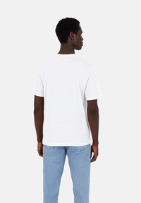 T-shirt in cotone bianco con maniche corte, collo rotondo e vestibilità rilassata. Indossato con jeans in denim blu chiaro. La vista posteriore mostra un design semplice.