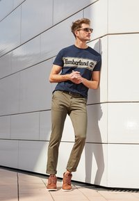 Homme portant des lunettes de soleil, un t-shirt Timberland bleu marine, un pantalon olive et des baskets marron, appuyé contre un mur carrelé blanc en extérieur.