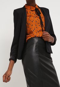 Blazer nero su una blusa arancione a fantasia con colletto e fiocco, abbinato a una gonna nera in pelle aderente. Trame lisce, design sartoriale.