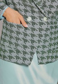 Grüne und weiße gemusterte Blazer mit einer gewebten Textur, ausgestattet mit einem Doppelknopfverschluss und einer Seitentasche. Seidiger mintfarbener Unterskirt sichtbar.